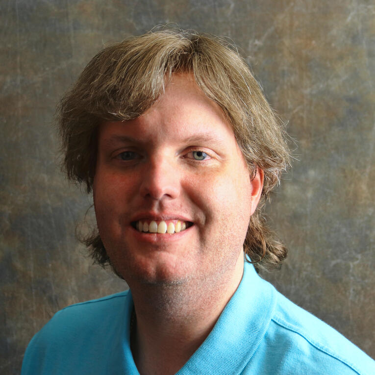 My headshot. I am standing against a tan matte and wearing a sky blue collared polo shirt.