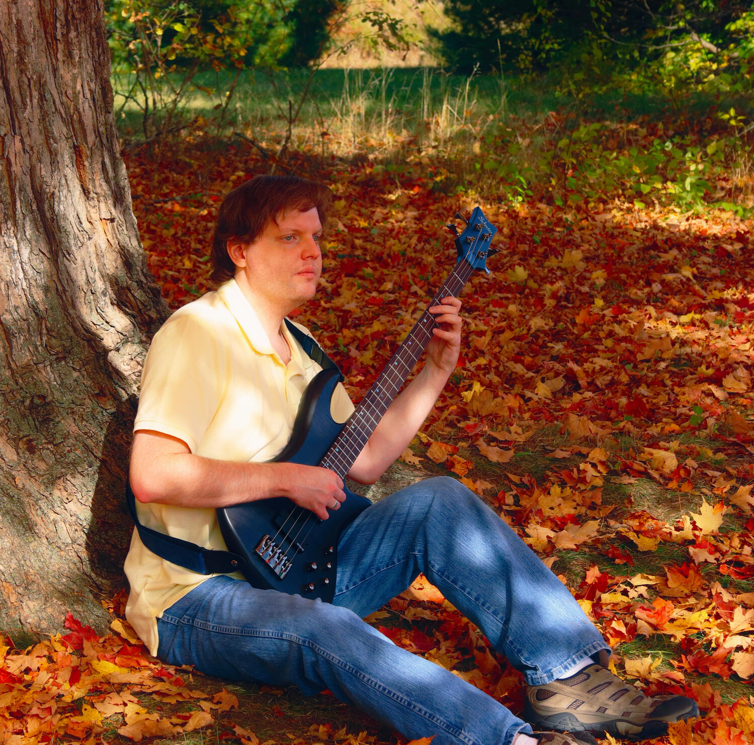 I am sitting beneath an oak tree in autumn in a bed of red, orange and yellow leaves. I am wearing a yellow polo shirt and jeans with a piccolo bass guitar in my lap, in playing position.